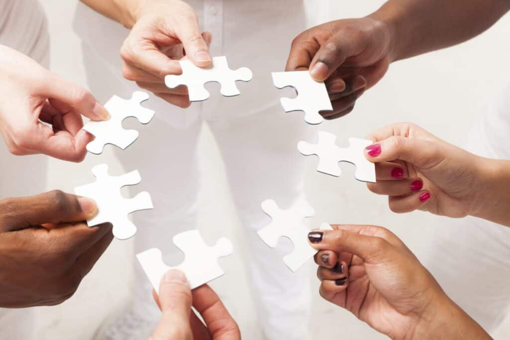 Six hands positioned in a circle all holding a single piece of a white puzzle