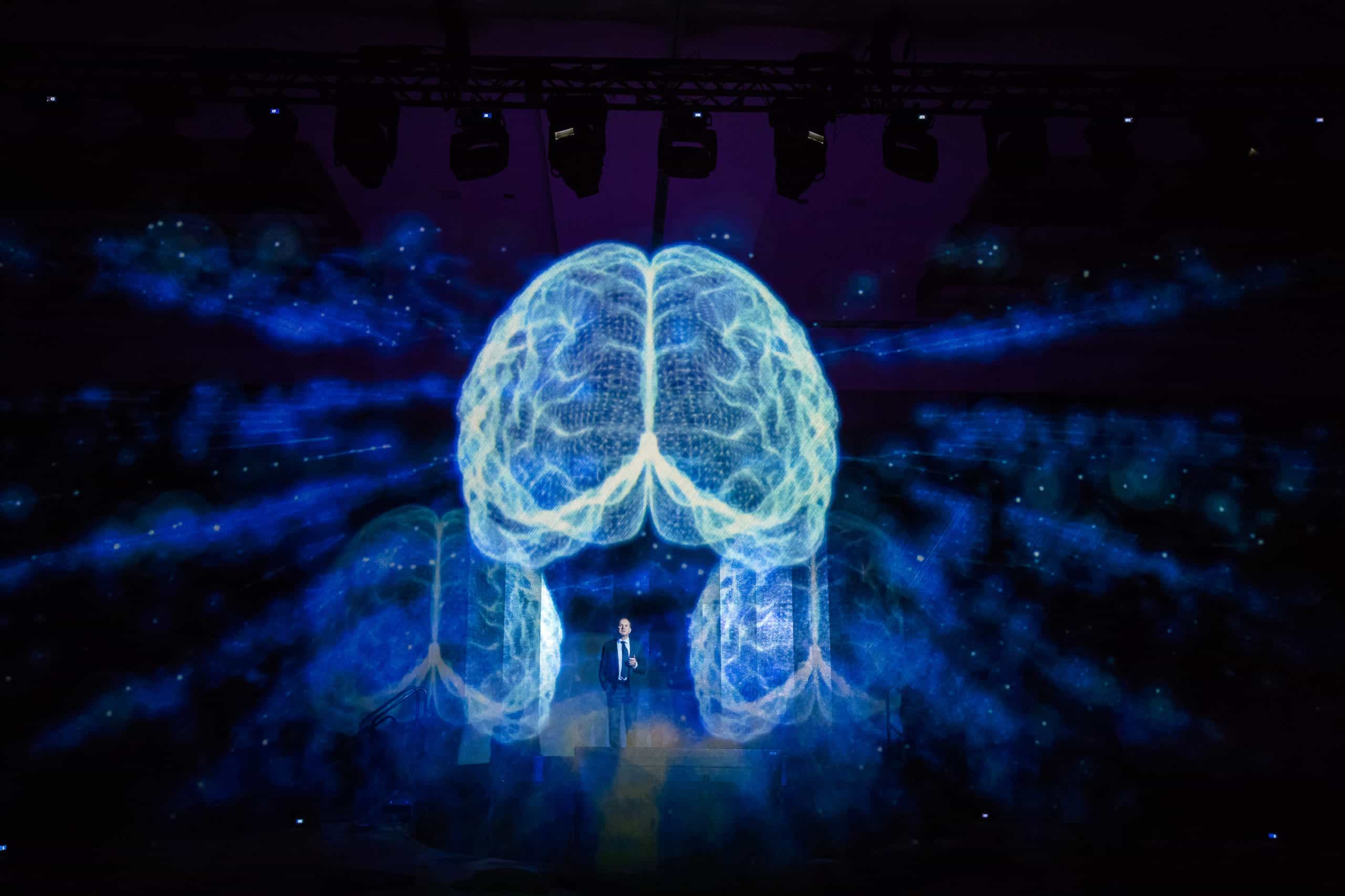 Speaker stands underneath a semi-transparent hologram of a blue human brain at a pharmaceutical industry conference