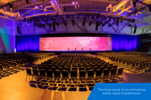 An empty conference room is filled with black chairs that facd a large stage with a large horizontal led screen.