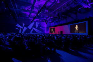 A dark room filled with attendees watching an individual woman speak on a stage at a corporate event.