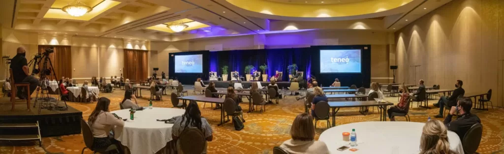 Panoramic view of Teneo corporate internal meeting, which shows several attendees sitting at round tables in front of a stage with projector screens on either side.