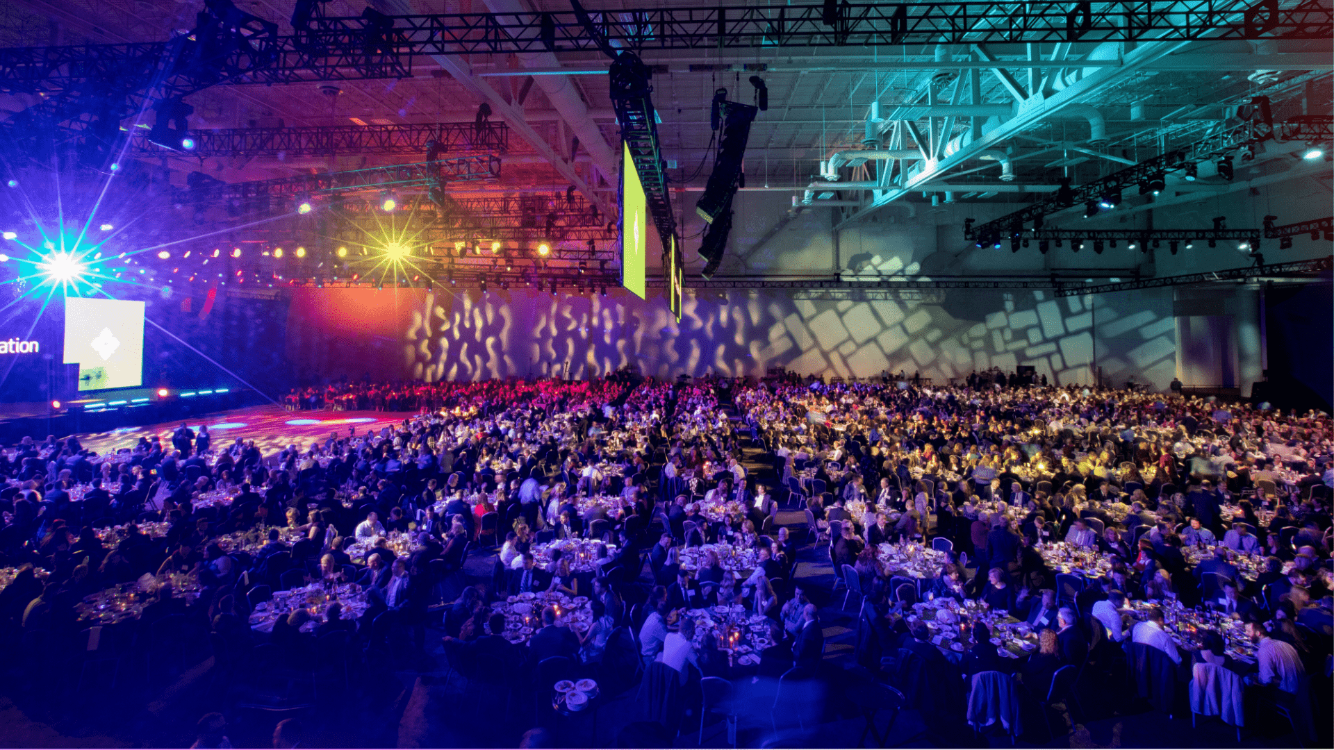 Large event space with creative lighting shining down on the attendees sitting at tables and looking at the stage