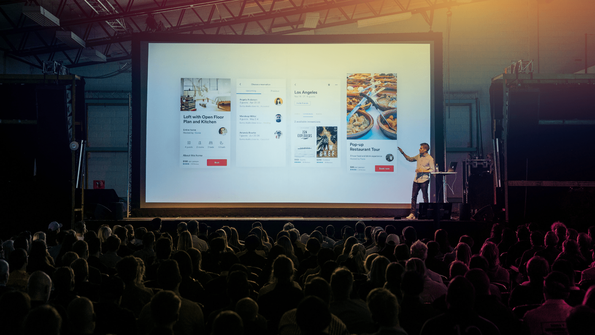 Man speaking and pointing to a very large screen on stage while the audience watching his presentation