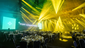LED lighting fills a corporate event room filled with round tables, a stage, and  multiple LED walls.